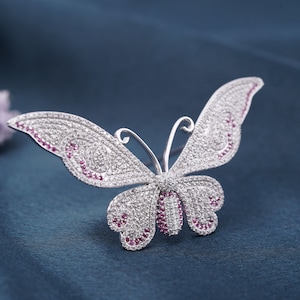 May include: A silver-toned butterfly brooch, encrusted with clear and ruby-colored crystals. The wings are detailed with intricate patterns, and the brooch is set against a dark blue background. This jewelry piece is a decorative accessory.