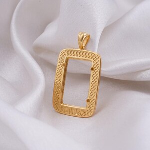 May include: A gold-colored rectangular pendant with a textured, open center. The pendant has a small loop at the top for a chain and is set against a white, silky background. The pendant's design features a detailed, woven pattern.
