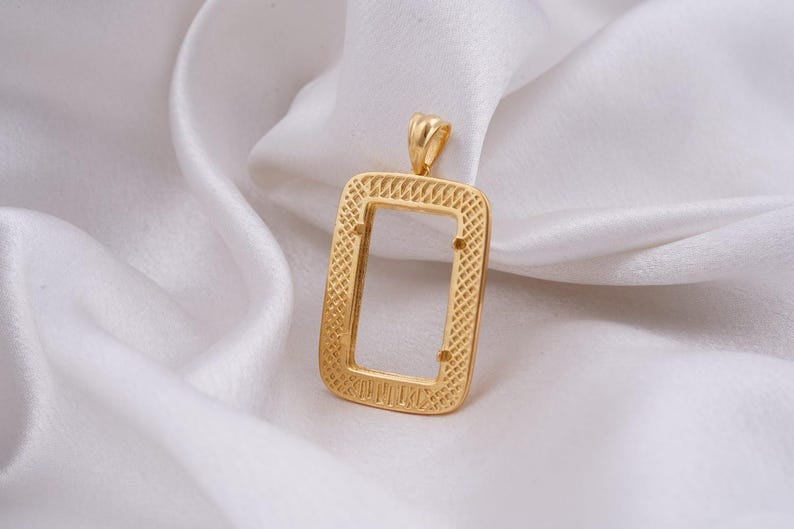 May include: A gold-colored rectangular pendant with a textured, open center. The pendant has a small loop at the top for a chain and is set against a white, silky background. The pendant's design features a detailed, woven pattern.