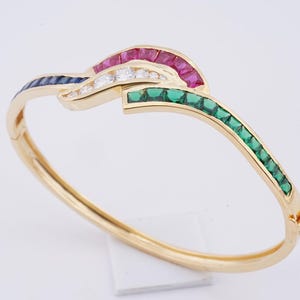 May include: A gold bangle bracelet with a colorful design. The bracelet features a curved design with sections of red, white, green, and blue gemstones. The bracelet is made of gold and has a clasp closure.