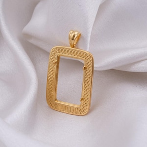 May include: A gold-colored rectangular pendant with a textured frame and a loop at the top. The pendant is displayed on a white, silky fabric, creating a contrast of textures and colors. The design is simple yet elegant.