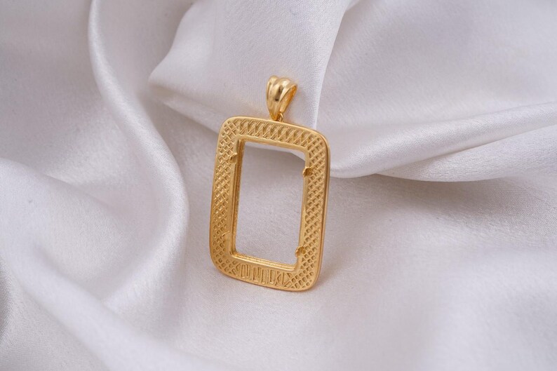 May include: A gold-colored rectangular pendant with a textured frame and a loop at the top. The pendant is displayed on a white, silky fabric, creating a contrast of textures and colors. The design is simple yet elegant.