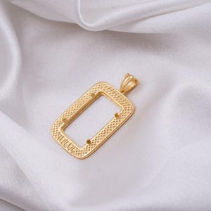 May include: A gold-colored rectangular pendant with a textured pattern and a small loop at the top. The pendant is displayed on a white, silky fabric.