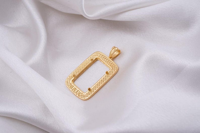 May include: A gold-colored rectangular pendant with a textured pattern and a small loop at the top. The pendant is displayed on a white, silky fabric.