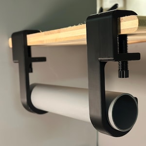 Könnte beinhalten: Schwarze Metallklemme, die an einer Holzoberfläche befestigt ist und eine weiße Röhre hält. Die Klemme hat eine Schraube zum Festziehen.