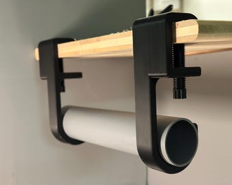 May include: Black metal clamp mounted to a wooden surface, holding a white tube. The clamp has a screw to tighten it.