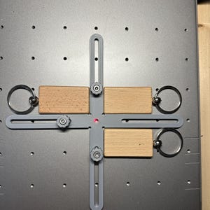 Könnte beinhalten: Vier Holzschlüsselanhänger mit Metallringen sind an einem grauen Metall-Jig mit einem roten Laserpunkt in der Mitte befestigt. Der Jig hat zwei verstellbare Knöpfe auf jeder Seite.