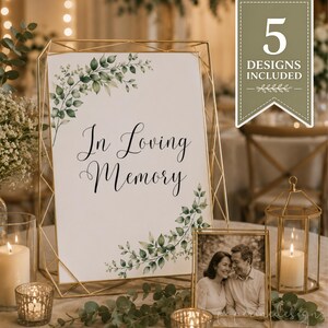 May include: A white rectangular sign with the black text In Loving Memory, decorated with green watercolor eucalyptus leaves in the corners. The sign is displayed in a gold geometric wire frame, accompanied by a framed photograph and candles.