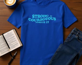 May include: A royal blue t-shirt with the text "Strong & Courageous Joshua 1:9" in light blue. The shirt is paired with blue jeans, a cup of coffee, a book, and a pen on a wooden surface.