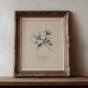 以下が含まれることがあります： 花とつぼみの白黒の植物画が描かれた額入りのアートプリント。プリントには「The joy of the Lord is my strength」というテキストが含まれています。フレームは金色の装飾が施されています。木製のテーブルの上にあります。