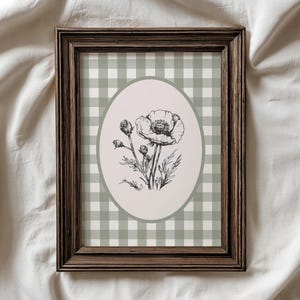 May include: A framed floral print featuring a black and white botanical illustration of a poppy flower. The artwork is set against a light green and white checkered background within a dark brown wooden frame. The print is displayed on a white fabric surface.