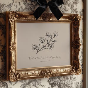 Puede incluir: Un marco dorado adornado con un lazo negro. La impresión presenta una ilustración floral y el texto "Trust in the Lord with all your heart". El marco está colgado en un papel pintado floral.