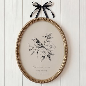Puede incluir: Una impresión enmarcada ovalada con un borde dorado con cuentas, que representa una ilustración en blanco y negro de un pájaro y flores. La impresión está suspendida por una cinta negra. El texto dice: "Sus misericordias son nuevas cada mañana, Lamentaciones 3:23".