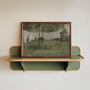 May include: A framed print of a vintage painting depicting white laundry hanging on a clothesline in a green field. The print is displayed on a green and tan shelf. The text on the print reads: "Whatever you do, in word or deed, do everything in the name of the Lord Jesus, giving thanks to God the Father through Him."
