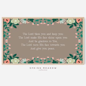 May include: A framed print with a floral border. The text reads, "The Lord bless you and keep you...And give you peace." The floral border features green leaves and pink, white, and peach flowers. The background is a muted brown.