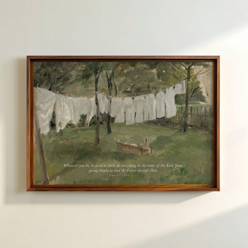 May include: A framed print of a pastoral scene. White laundry hangs on a clothesline in a green field. The artwork has a brown wooden frame and includes text at the bottom: "Whatever you do, in word or deed, do everything in the name of the Lord Jesus, giving thanks to God the Father through Him."