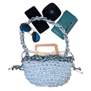 May include: A light blue woven handbag with a wooden handle and a braided strap. The bag is filled with a cream-colored lining. Sunglasses, a teal wallet, and two black cases are also in the image.