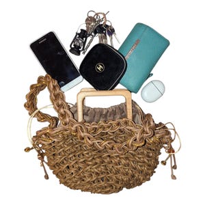 May include: A woven brown handbag with a wooden handle, filled with everyday items. Includes a smartphone, keys, a black case with a logo, a teal wallet, and a white wireless earbud case.