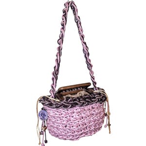 May include: A woven pink handbag with a braided handle in shades of pink and black. The bag has a decorative trim and a small charm. Inside the bag is a wooden object.