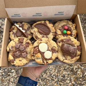 May include: A selection of six chocolate chip cookies in a cardboard box, each decorated with various chocolate toppings. The box is labelled "SWEET ADDICTION". The cookies are held in a person's hand.