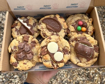 Puede incluir: Una caja de seis galletas con chispas de chocolate, cada una cubierta con diferentes chocolates. Las galletas están en una caja de cartón con el texto "SWEET ADDICTION". La caja es sostenida por una persona.