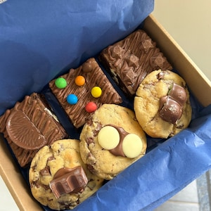 May include: A selection of baked treats in a box, including brownies and cookies. The brownies are decorated with chocolate and sweets. The cookies feature chocolate chips and candy. The box is lined with blue tissue paper.