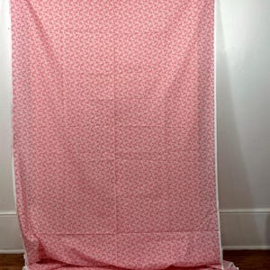 Puede incluir: Un gran trozo de tela rosa con un patrón repetitivo de pequeñas formas rosa claro. La tela está drapeada, con pliegues y arrugas visibles. Los bordes están acabados con un borde blanco. La tela probablemente se utiliza para coser o manualidades.