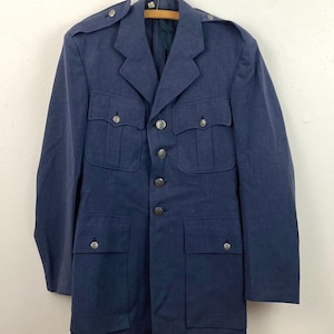 May include: A vintage blue jacket with four front pockets, shoulder epaulets, and silver-toned buttons. The jacket is hanging on a wooden hanger against a white background. The jacket has a classic military style.