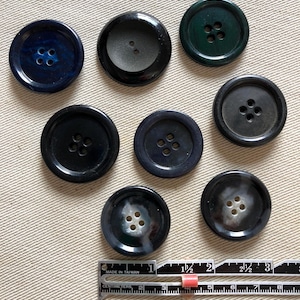 May include: A collection of eight round buttons in various shades of blue, green, and black. The buttons have four holes and are arranged on a beige surface. A ruler with both inches and centimeters is at the bottom.