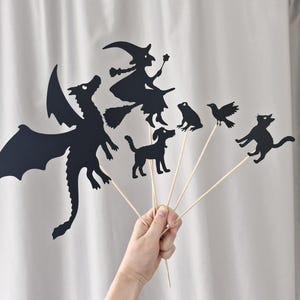 May include: A set of black paper silhouettes attached to wooden sticks, featuring a dragon, a witch on a broomstick, a dog, a frog, a bird, and a cat. These decorative cutouts are arranged in a fan shape, showcasing detailed outlines of each character against a plain background.