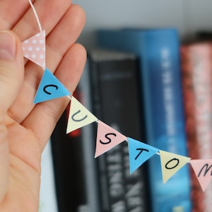 May include: A paper banner with the word "CUSTOM" spelled out in colorful triangular flags. The flags are pink, blue, and yellow with white polka dots. The banner is held in a hand against a blurred background of books.