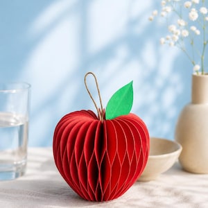 Könnte beinhalten: Eine rote Apfeldekoration aus Wabenpapier mit einem grünen Papierblatt und einer dünnen braunen Schnurschlaufe an der Oberseite. Der Apfel hat eine segmentierte Ziehharmonika-Struktur, die eine abgerundete, dreidimensionale Form erzeugt.