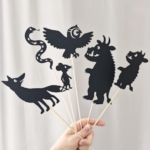 May include: A set of six black paper silhouettes attached to wooden sticks, featuring a fox, a snake, an owl, a mouse, a horned monster, and a small creature. Each cutout is detailed with white eye accents and distinct character shapes.