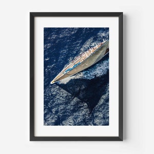 Könnte beinhalten: Eine Luftaufnahme eines großen weißen Segelboots mit einem blauen Streifen, das auf einem tiefblauen Meer segelt. Das Boot ist von Gischt umgeben und die Sonne spiegelt sich im Wasser.