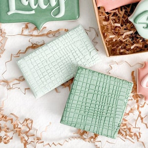 May include: A collection of decorated cookies with names written on them, including Lucy, Maddie, Sophie, and Sammie. The cookies are arranged in a box with brown paper shredding. There are also two plain cookies with a green and white checkered pattern.