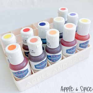 May include: A white plastic container with the words "Apple & Spice" and "Custom Cookies" printed on it. The container holds 15 small bottles of food coloring, each with a different color. The bottles are labeled "AmeriColor" and have a lot number printed on them.