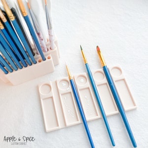 May include: A set of four blue paint brushes with gold tips in a white plastic holder with five wells. The holder has a white paint splatter on it.