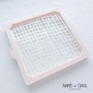 May include: A light pink cookie stencil with a square shape and a white, abstract pattern. The stencil has rounded corners and small, decorative tabs on two sides. The text "Apple & Spice Custom Cookies" is visible in the bottom right corner.