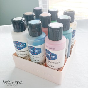 May include: A set of 10 bottles of AmeriColor food coloring in a pink plastic organizer. The bottles are labeled with the brand name and the color name. The colors include white, blue, teal, pink, green, and yellow.