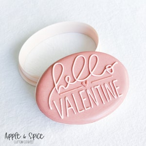 May include: A pink oval cookie with white icing that says "hello valentine" with a small pink heart in the middle of the word "hello".