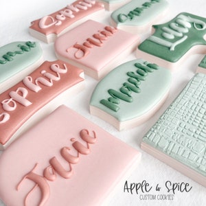May include: A collection of decorated sugar cookies with various names written in cursive on them. The cookies are in shades of pink, green, and brown. The cookies are arranged on a white surface. The text "Apple & Spice CUSTOM COOKIES" is visible at the bottom of the image.