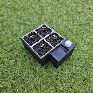 Op de afbeelding: Een zwart-witte zaailingstarterbak met vier vierkante compartimenten gevuld met aarde en kleine groene planten. Een waterreservoir en een witte dop zijn aan de zijkant bevestigd. De bak staat op een groene grasachtige ondergrond.