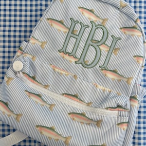 May include: A light blue and white striped backpack with a pattern of rainbow trout. The backpack has a front zippered pocket and a top handle. The initials "HBI" are embroidered in green on the front.