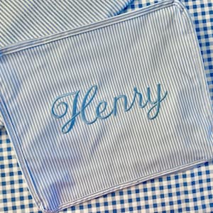 May include: A blue and white striped fabric item with the name "Henry" embroidered in a light blue script. The item is rectangular and appears to be a pillowcase or similar fabric accessory. The background is a blue and white gingham pattern.