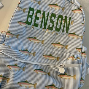 May include: A blue and white striped backpack with a repeating pattern of colorful fish. The name "BENSON" is embroidered in green on the front. The backpack has a zippered front pocket and adjustable straps.