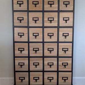 May include: A tall, wooden cabinet with twenty-four drawers. The drawers are light-colored wood with black metal label holders and pulls. The cabinet frame is black, and it sits on small, decorative feet.