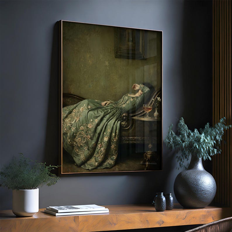 May include: A framed wall art print depicting a person in a long, flowing sage green gown with intricate gold floral embroidery, reclining on a dark curved sofa. A small side table holds a glass of amber liquid and a single white flower.