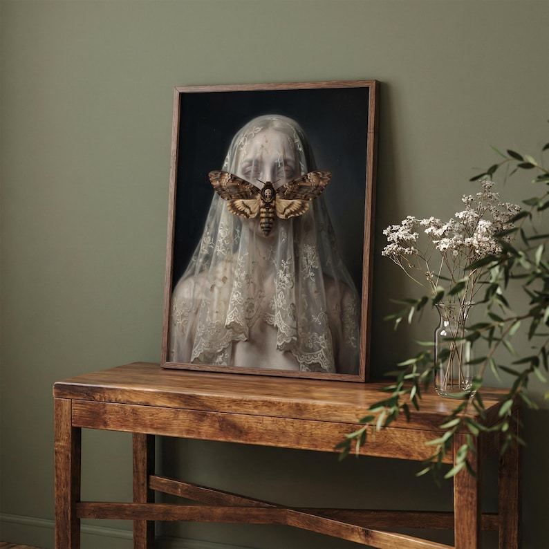 May include: Framed artwork featuring a person veiled in lace, with a large moth covering the face. The moth has brown and tan markings. The artwork is displayed on a wooden table against a dark background.