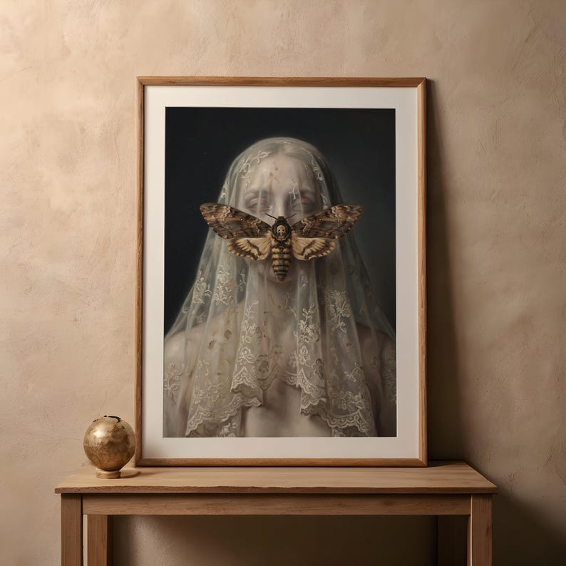 May include: Framed art print featuring a portrait of a person veiled in lace, with a large moth covering the face. The moth has intricate wing patterns in shades of brown and tan. The artwork is displayed on a wooden table.