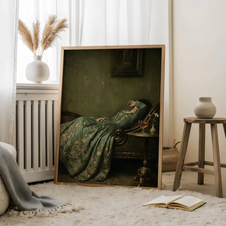 May include: A framed wall art print depicting a person in a long, flowing green dress with intricate floral embroidery, reclining on a dark ornate chaise lounge in a dimly lit room, with a small side table holding a glass and a tiny vase nearby.
